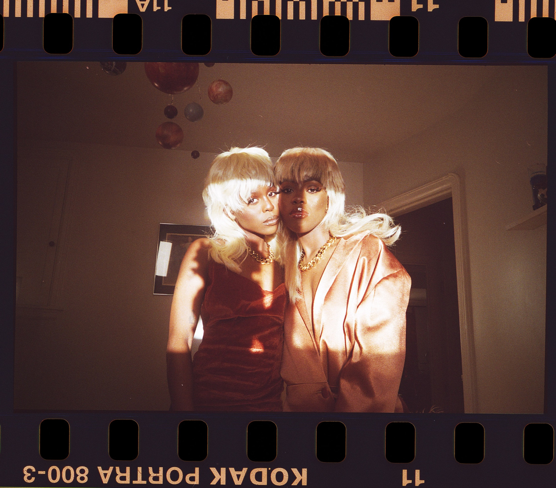 A photograph of two Black women and twin sisters, Kakra & Penny, embracing each other while dressed in matching blond mullets & gold chains. One wears a brown satin dress while the other wears an oversized champagne blazer; light shines brightly through the window on both of their faces as they look straight to camera.