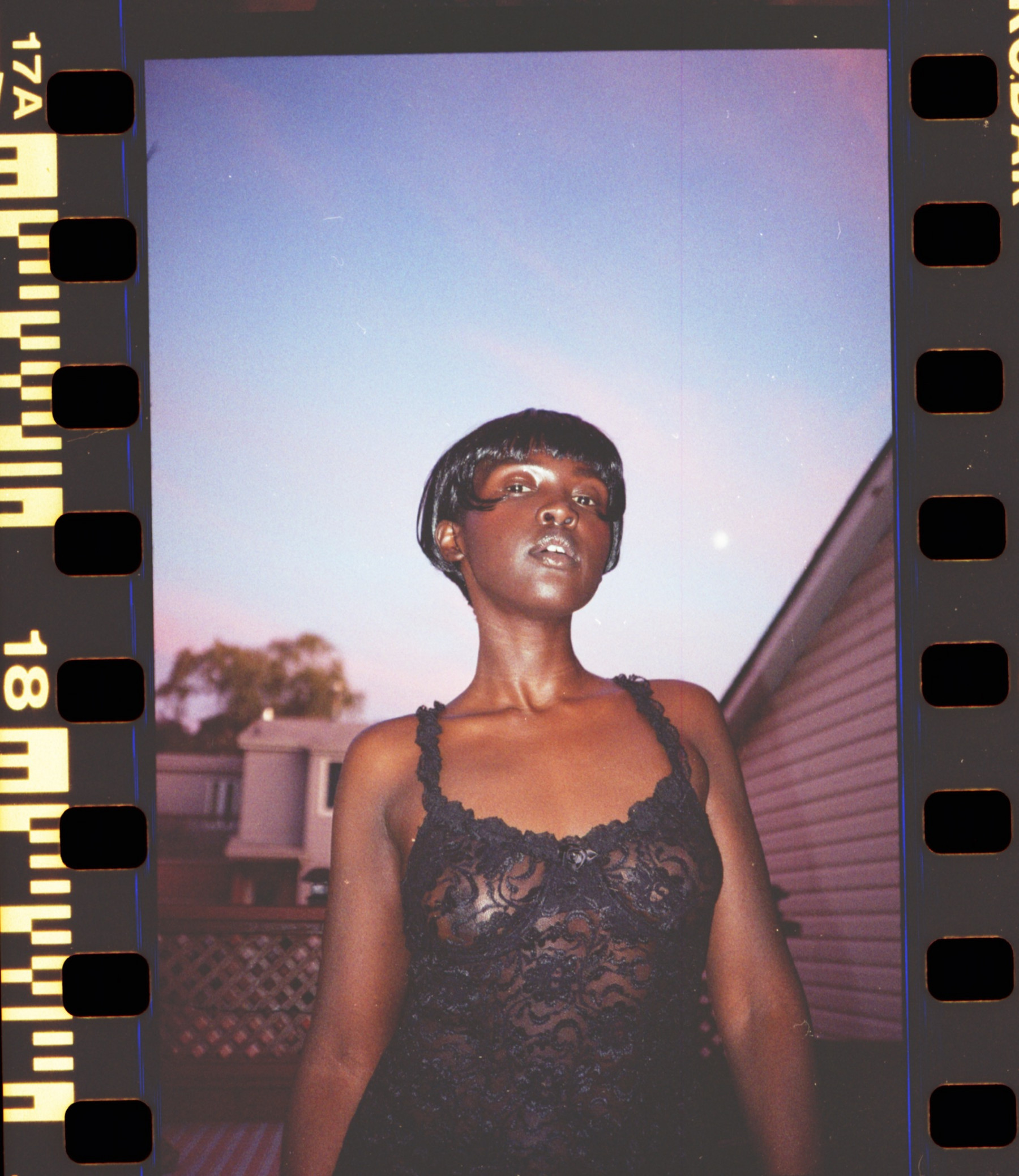 A Black woman whose hair is styled in a short bob stands on a rooftop at twilight, looking down to camera. Flash lights up her face, while the remnants of sunset light up the sky behind her.