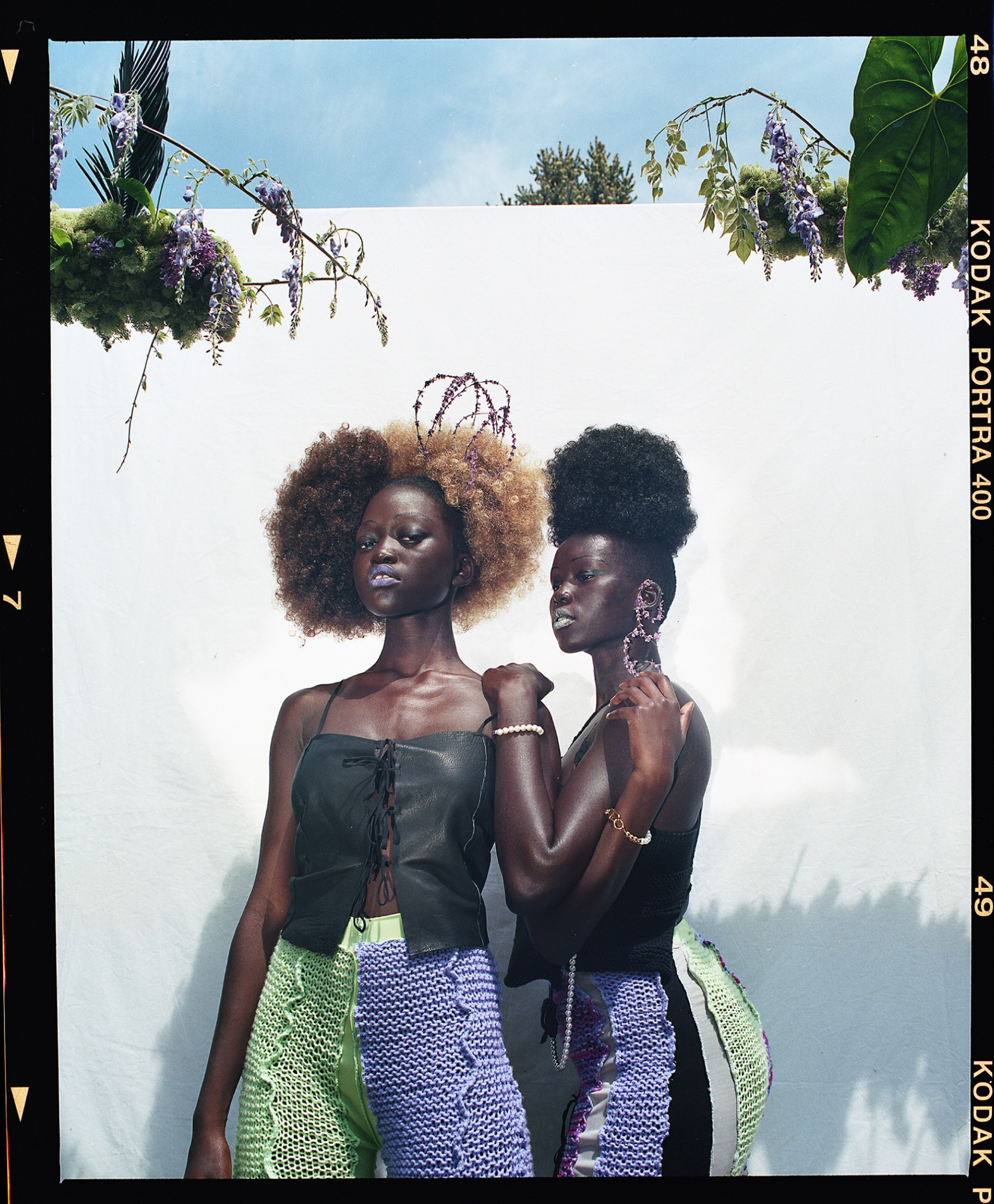 Two Black women stand outside on a sunny day in front of a white backdrop framed with florals as they hold each other and look directly to camera. They wear Black tops and matching purple, green and black crochet bottoms.