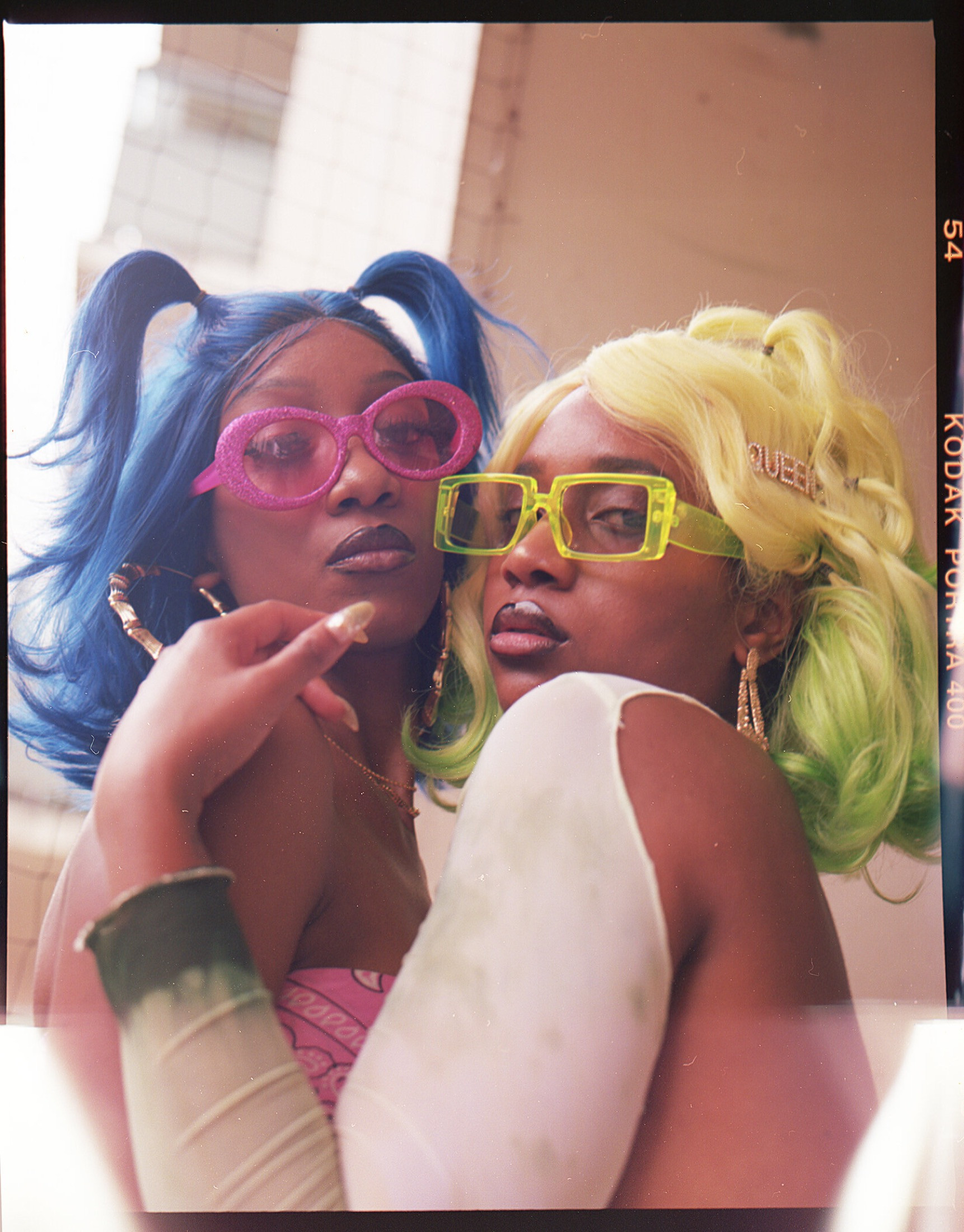 A close-up shot of two women (one wearing a pink top and pink sunglasses with blue hair; one wearing a cream shirt and neon yellow sunglasses with blond hair and green dip-dyed ends) hold each other and look down at the camera.
